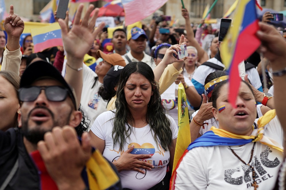 Woman in center of frame, surrounded by large crowd; Venezuelan flag colors on garments, demonstration items and flags. 