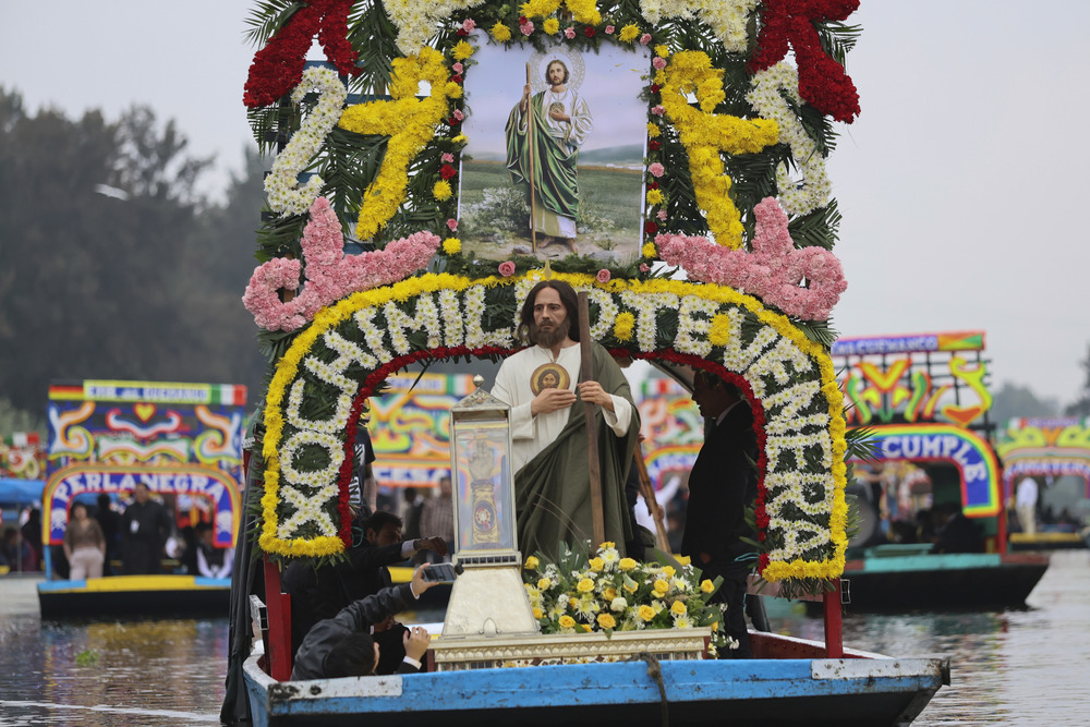 Man dressed as St. Jude Thaddeus, stands on front of decorated barge, floating down river and carrying a relic of St. Jude. 