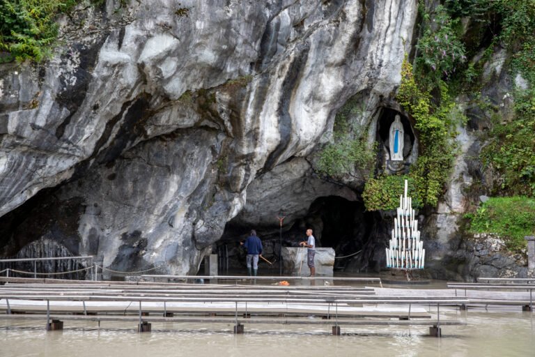 Statue of Our Lady of Lourdes nestled in side of cliff; flood-waters obscure gathering space. 