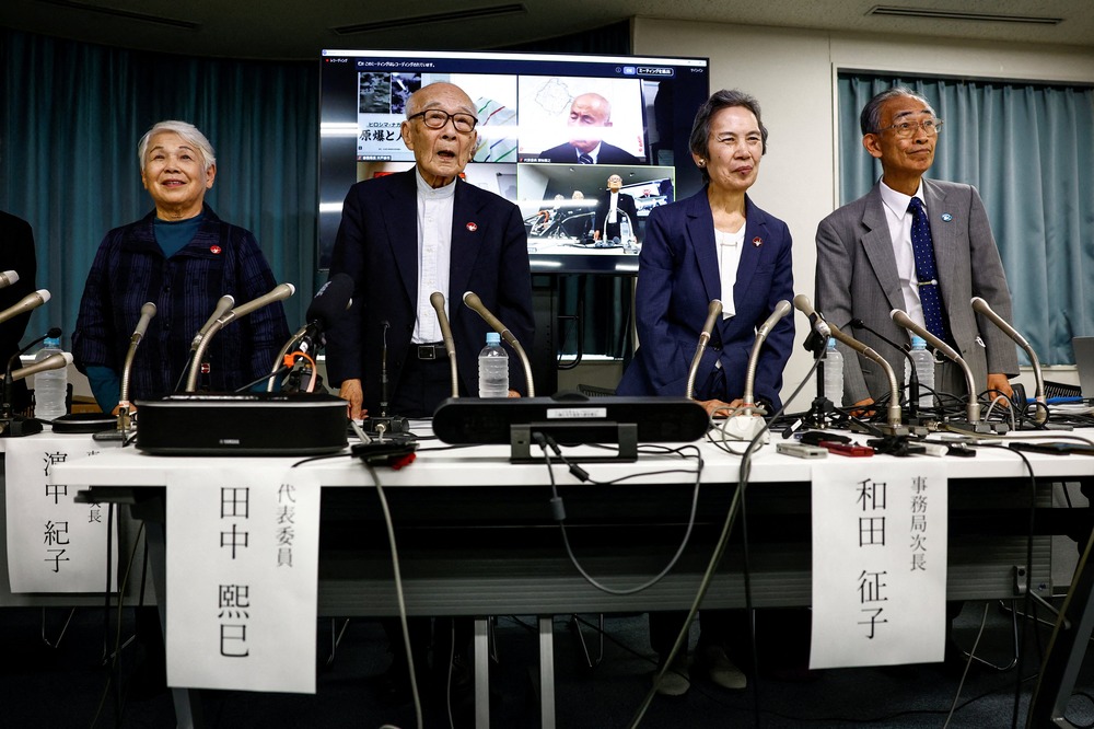 Four elderly people stand behind a table with microphones at press conference; behind is a screen.