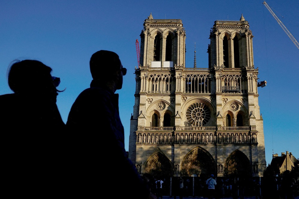 The Cathedral looms against blue sky; on right side is the arm of a crane; in left third of frame are two silhouetted figures of bystanders.