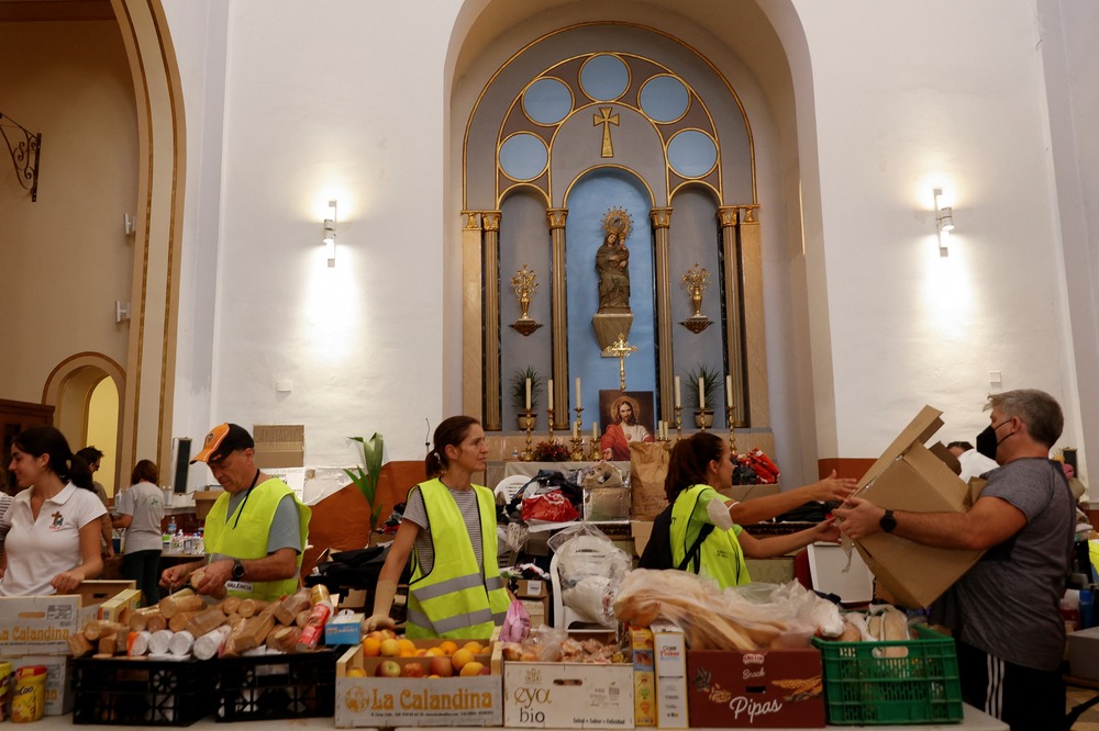 Volunteers wearing yellow reflective vests work behind long table covered with supplies; in the background are the windows and wall of old church.