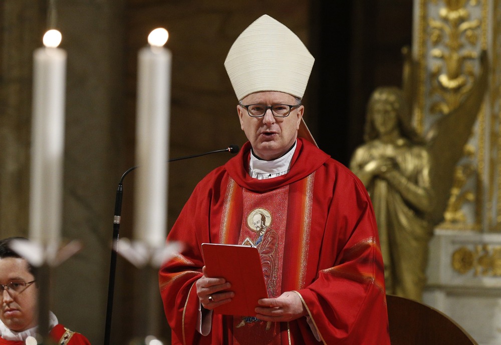 The bishop is vested for mass in red, wearing mitre, and delivering remarks.