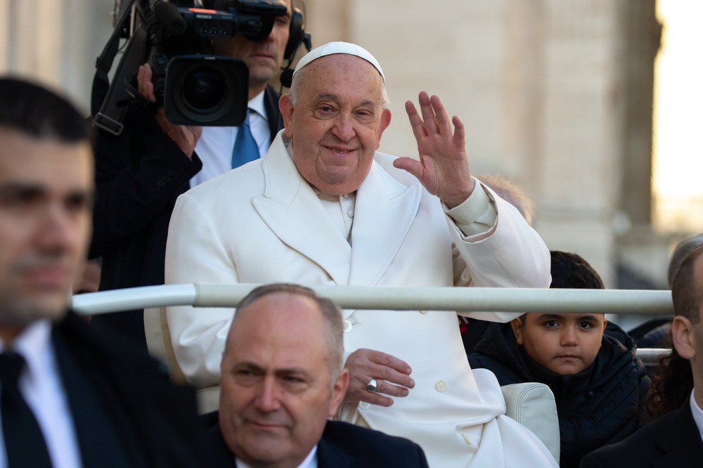 Francis seated, hand lifted in greeting and smiling.