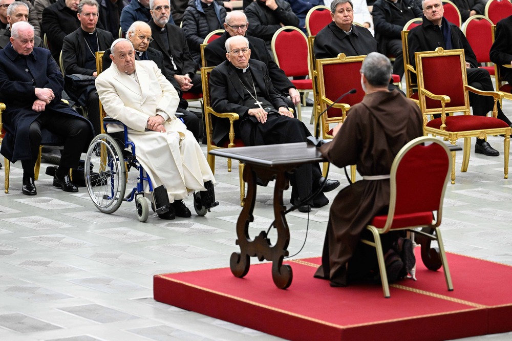 Pasolini sits on red dais, shown from behind, Facing his is Pope Francis in wheel chair, and many Cardinals clothed in black clerics and seated in red chairs.