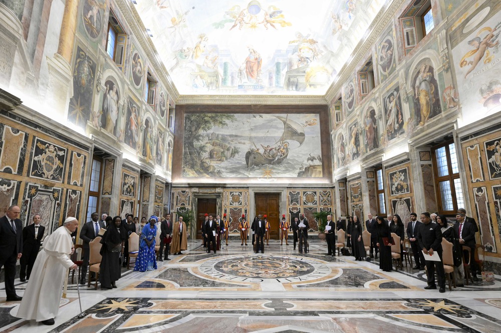 Large frescoed room; diplomats sit in u-shaped formation of chairs, Pope enters walking on cane. 