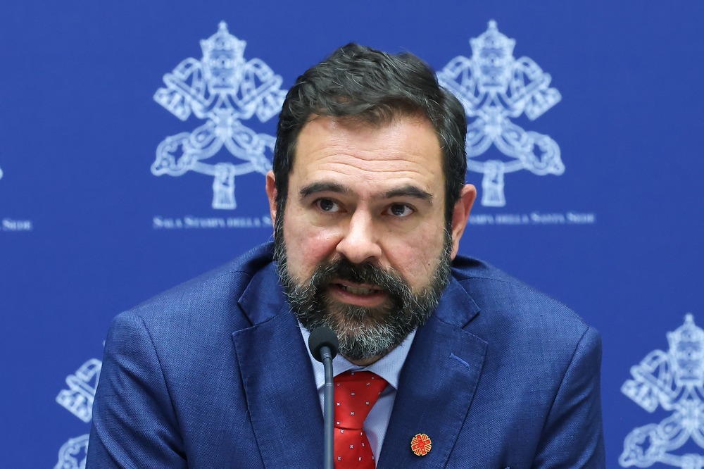 Cervantes pictured sitting at press conference wearing blue suit and red tie. 