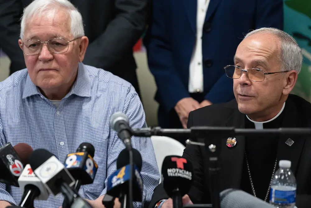 The two men sit side by side in front of microphones, Seitz wears black clerics.