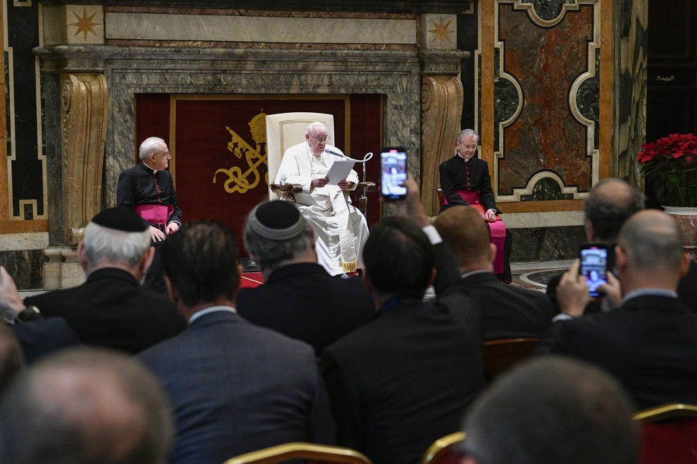 Pope sits on raised dais, people seen watching.