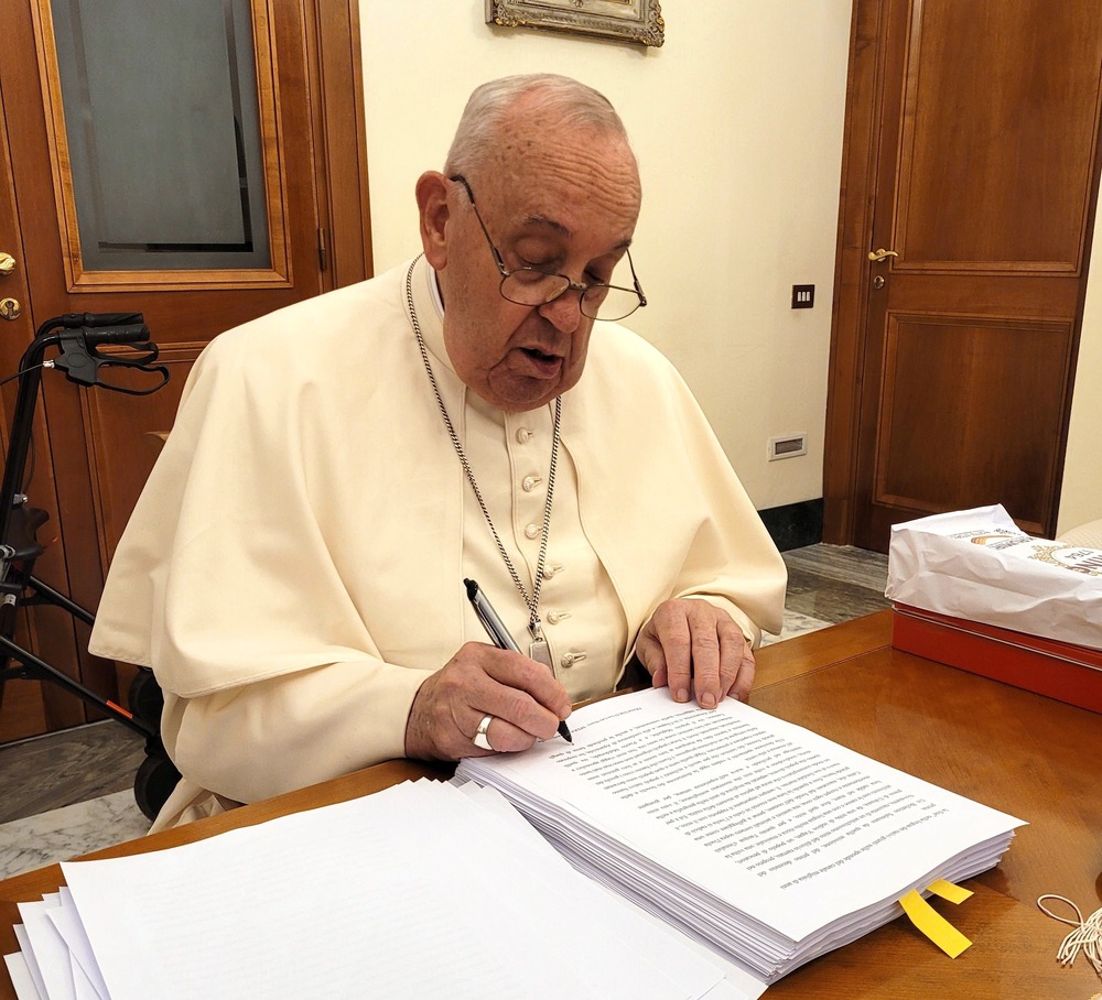 Francis sits at desk marking pages with pen. 