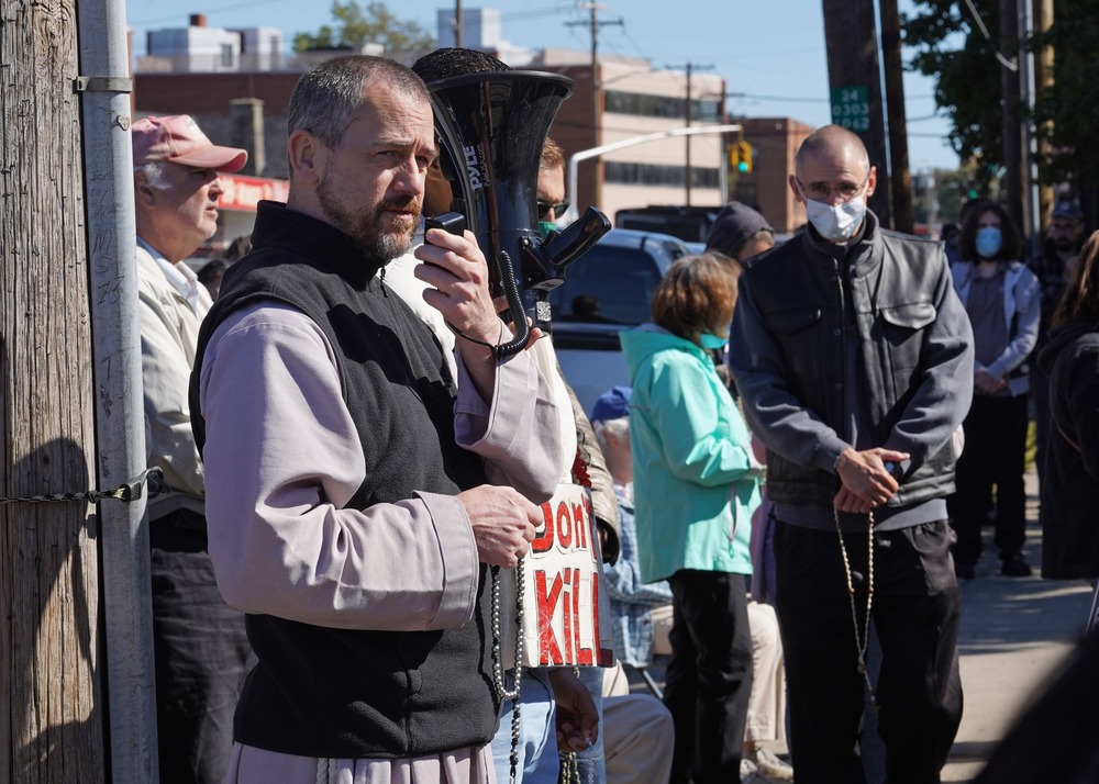 The priest stands holding megaphone and rosary.