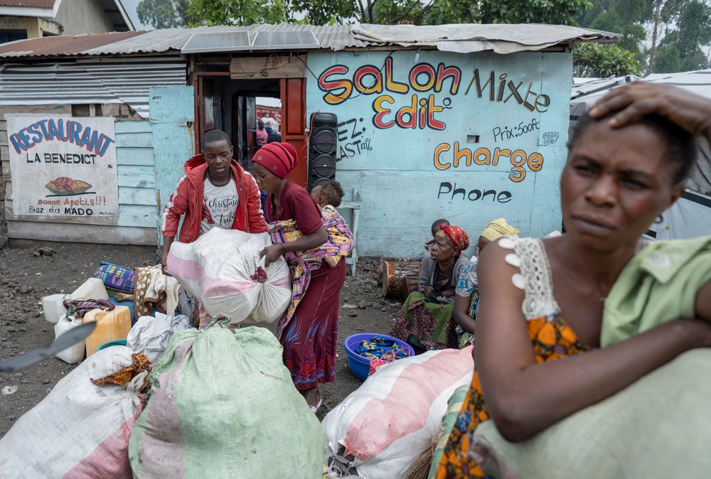 A woman is foregrounded leaning on sacks with hand on head, in background is a small storefront and and other people, including a woman with a baby on her back.