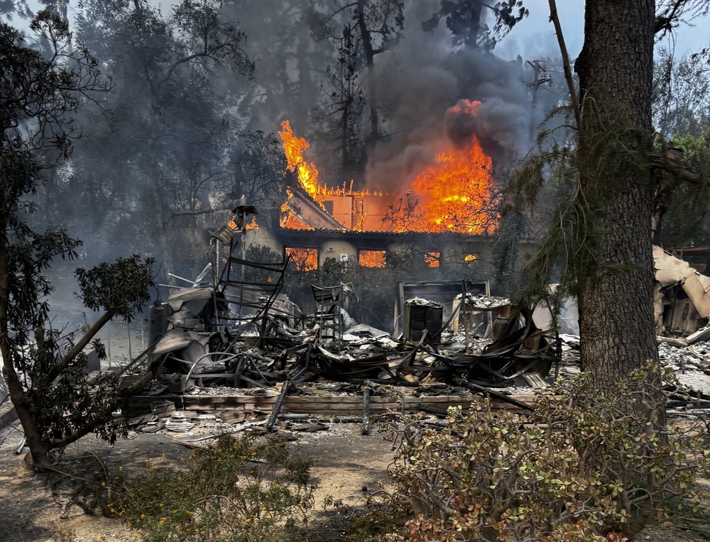 Fram filled by smoke and bordered by trees; centered is blackened shell of two-story house still burning.