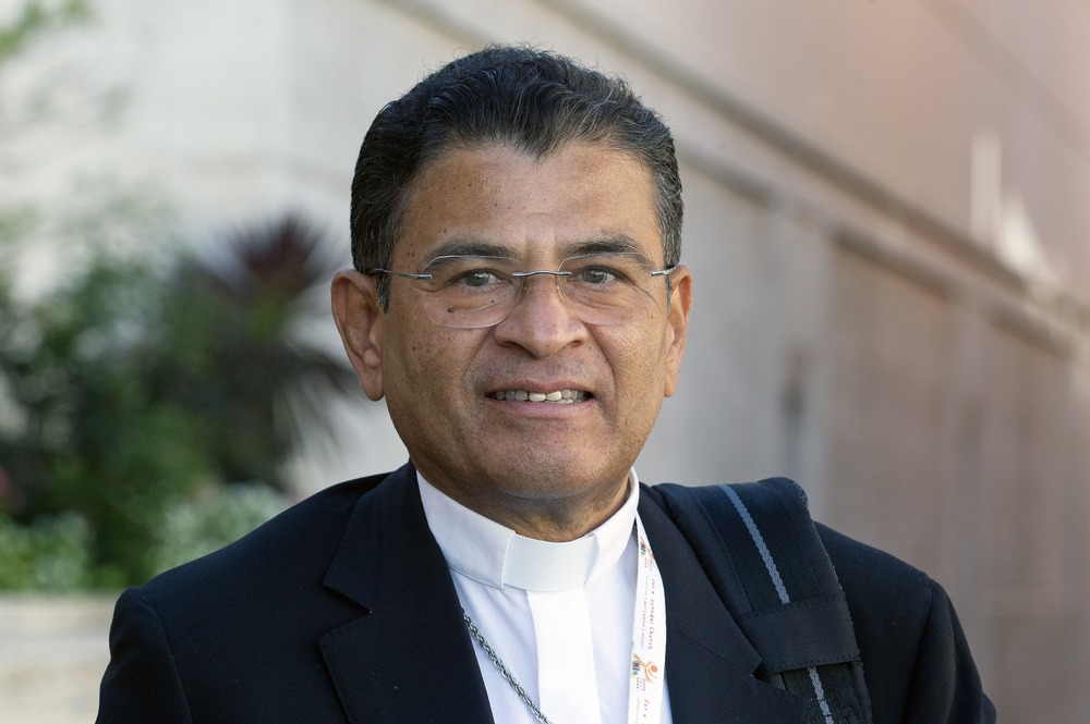 The bishops wears a black suit over white shirt with color, smiles at camera.