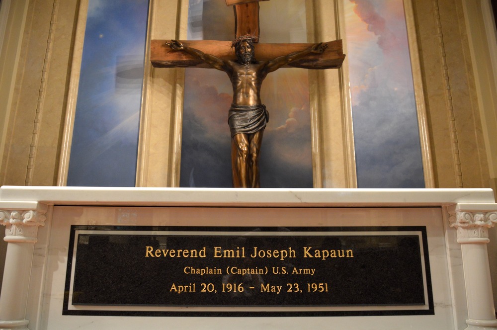 Large bronze crucifix hangs over marble tomb bearing plaque.