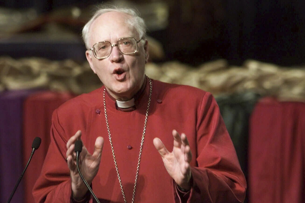 Carey shown with hands mid-gesture while speaking, wearing red cassock.