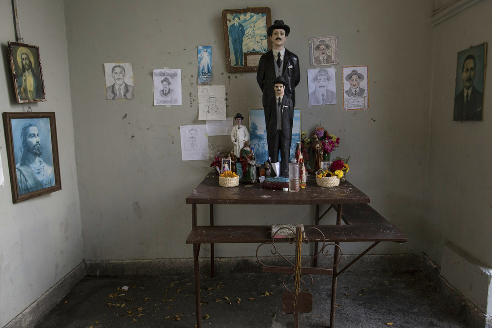 A large colored statue of Hernandez stands ont ables with flowers and other religious articles in a small gray room; walls covered with pictures and drawings of both Jesus Christ an Hernandez. 