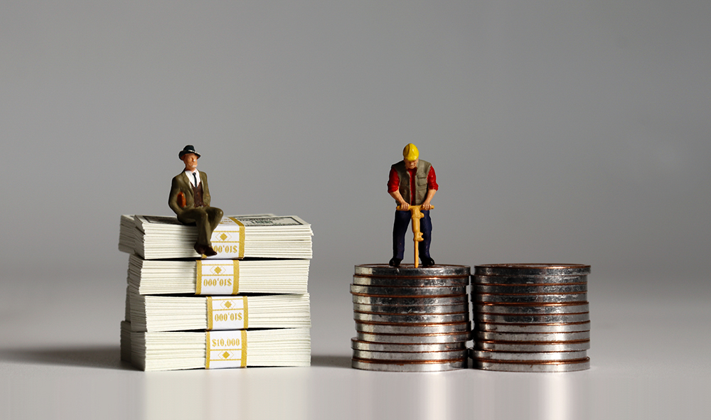 Figurine of a businessman sits on a pile of $100 bills while the figurine of a construction worker works on a shorter pile of coins (Dreamstime/Hye Jin Kang)