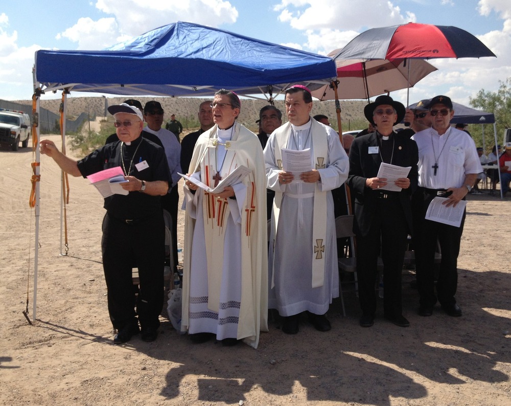 The archbishop wears white vestments and reads from book, others assembled around under tarpaulin tent and umbrellas.