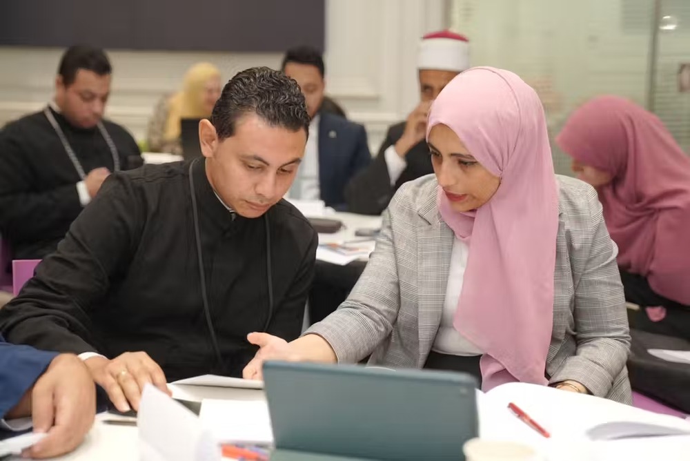 Benadi wearing business garb and pink hijab sits at a table with clergyman explaining concepts.