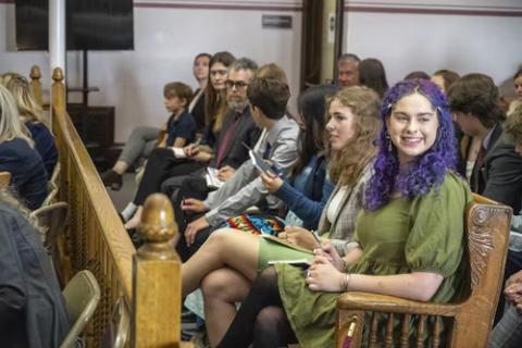 Rows of young people sit in courtroom benches; one smile in direction of camera. 