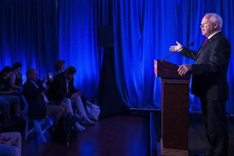 Gov. Walz pictured in profile, standing at lectern speaking with right hand raised in gesture. Room is covered in blue pipe and drape. 