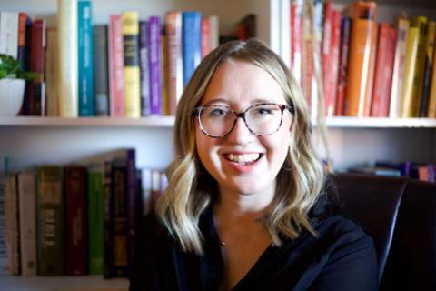 Thiessen smiles from desk chair, in background is bookshelf. 