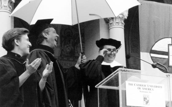 Black and white photo of the three gathered under umbrella in front of lectern. 