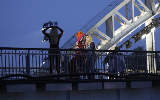 Two queens stand on bridge, dimly lit from behind, silhouetted camera operator visible in foreground. 