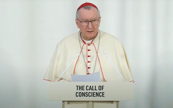 Parolin wearing white cassock piped with Cardinal's red and red zucchetto stands at podium reading remarks. 