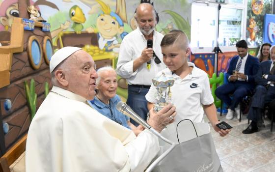 Pope Francis, seated next Sister Jeanningros, holds carousel and smiles; young boy who presented gift stands in front of Francis, watching his reaction. 