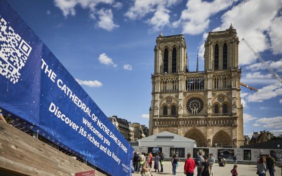 Notre Dame de Paris seen in background against bright blue sky; foregrounded is a tarp banner announcing the reconstruction and reopening of the Cathedral. 