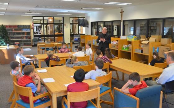 Children sit at table in large room, while priest stands talking. 