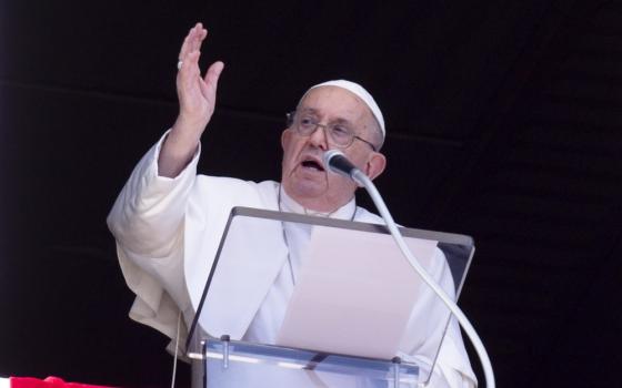 Francis stands in window of Apostolic Palace, in front of lectern, hand raised in blessing. 