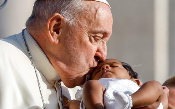 Close-up, square photo of Pope kissing a sleeping infant in the warm glow of the sun. 