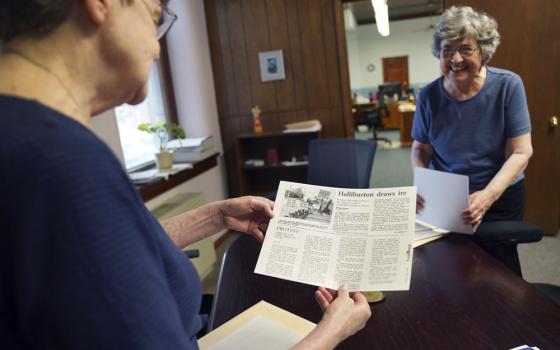 The two sisters, wearing plainclothes, stand in wood-paneled office, looking at clippings and smiling. 