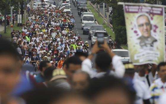 Large banner of Romero carried above long assembly of people marching down street. 