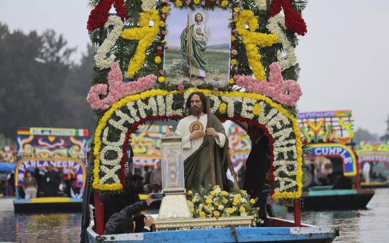 Man dressed as St. Jude Thaddeus, stands on front of decorated barge, floating down river and carrying a relic of St. Jude. 