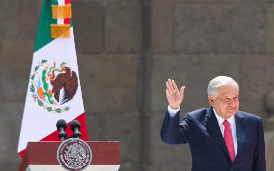 Obrador raises hand in greeting, standing on a dais; behind him is a podium and a Mexican flag. 