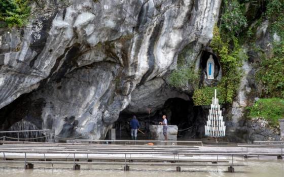 Statue of Our Lady of Lourdes nestled in side of cliff; flood-waters obscure gathering space. 