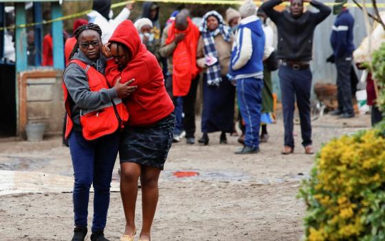 A crowd of mourners; two women walk away towards the camera, one holds the other as he cries. 