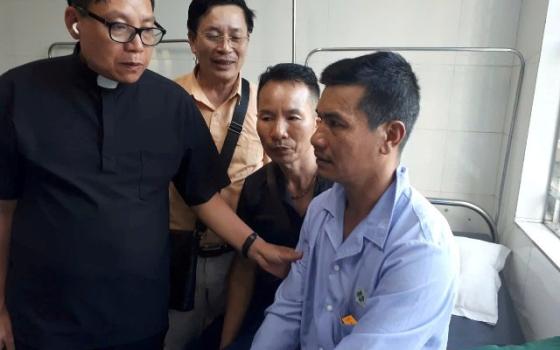 A man sits on hospital bed; next to him are the priest and two others.