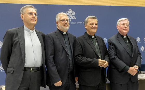 The four men stand in front of a blue and white photo background marked with the synodal insignia.