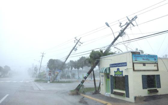 Windswept, rainy view of utility poles fallen; one leans on small building.