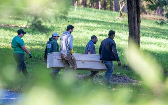 Seen through a frame of leaves, are five people, four of whom carry a casket.