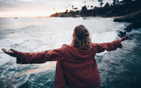 A woman stands with her arms outstretched, facing a coast in this photo illustration. (Unsplash/Nathan McBride)