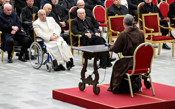 Pasolini sits on red dais, shown from behind, Facing his is Pope Francis in wheel chair, and many Cardinals clothed in black clerics and seated in red chairs.