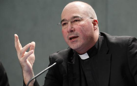 The priest wears black clerics and speaks and gestures into microphone.