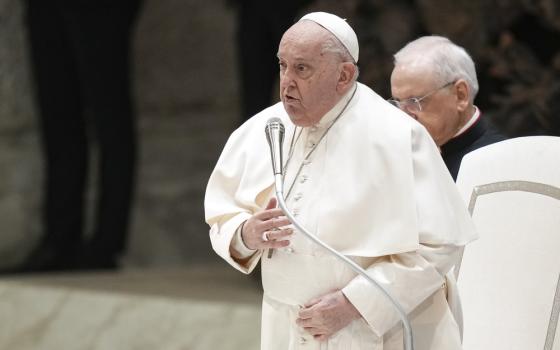 The pope stands speaking into microphone with hand to chest.