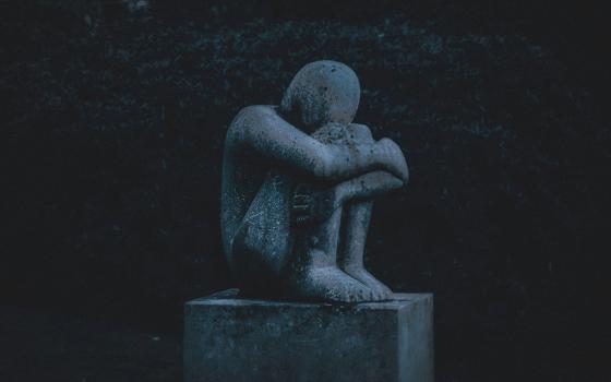 A sculpture depicts a crouched figure holding themselves, their head burrowed against their knees, arms crossed around their legs, in a dark outside setting (Unsplash/K. Mitch Hodge)